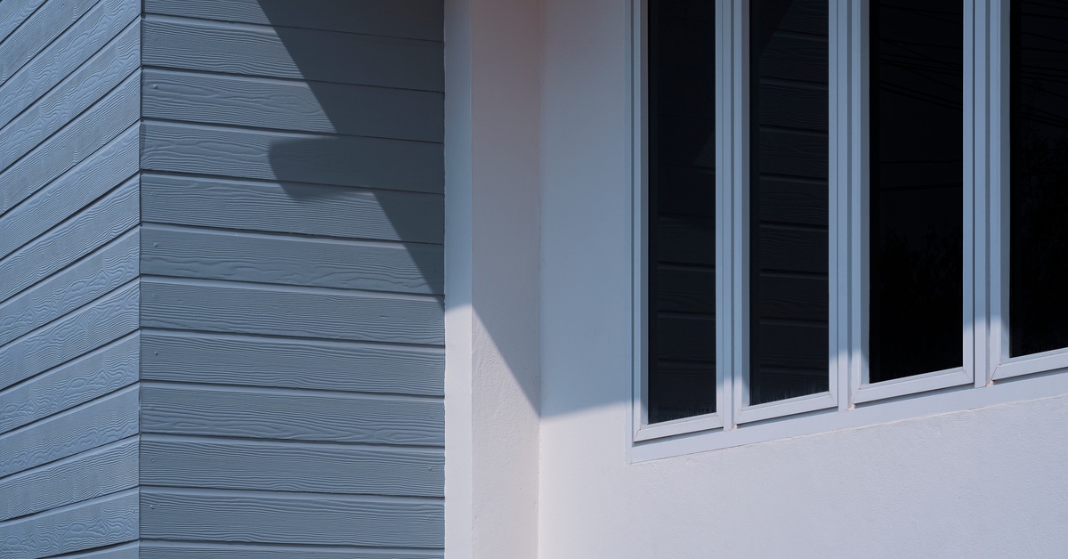 Four thin and long windows with tint placed on a white wall with a roof casting a shadow over it next to a blue wall.
