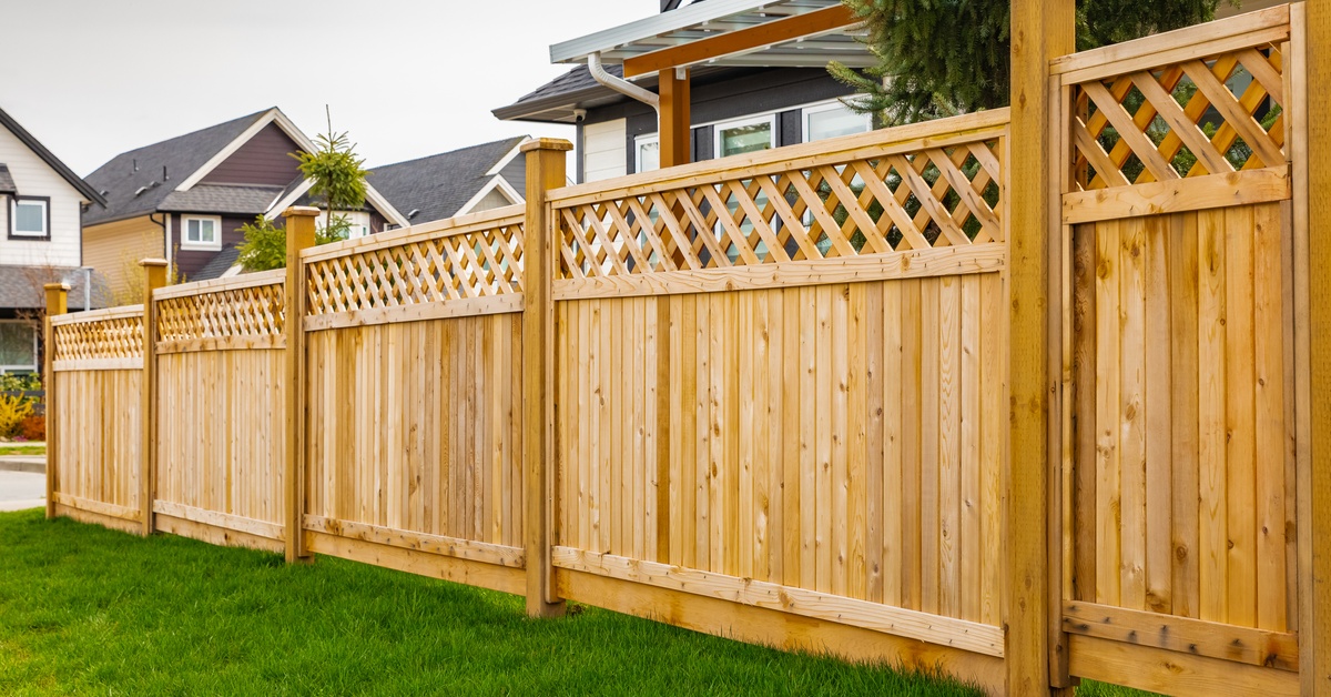 A wooden fence with a detailed top placed on the edge of a front yard over green grass with a house in the background.