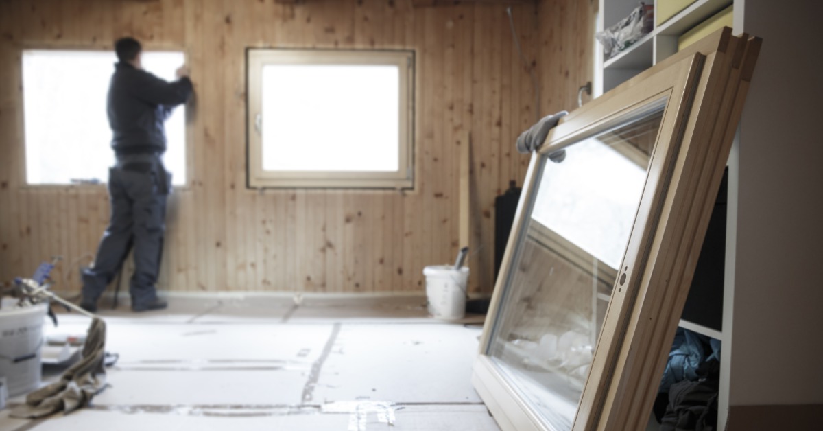 A worker sealing the edge of a window in a room under construction with a large double panel window on the floor.