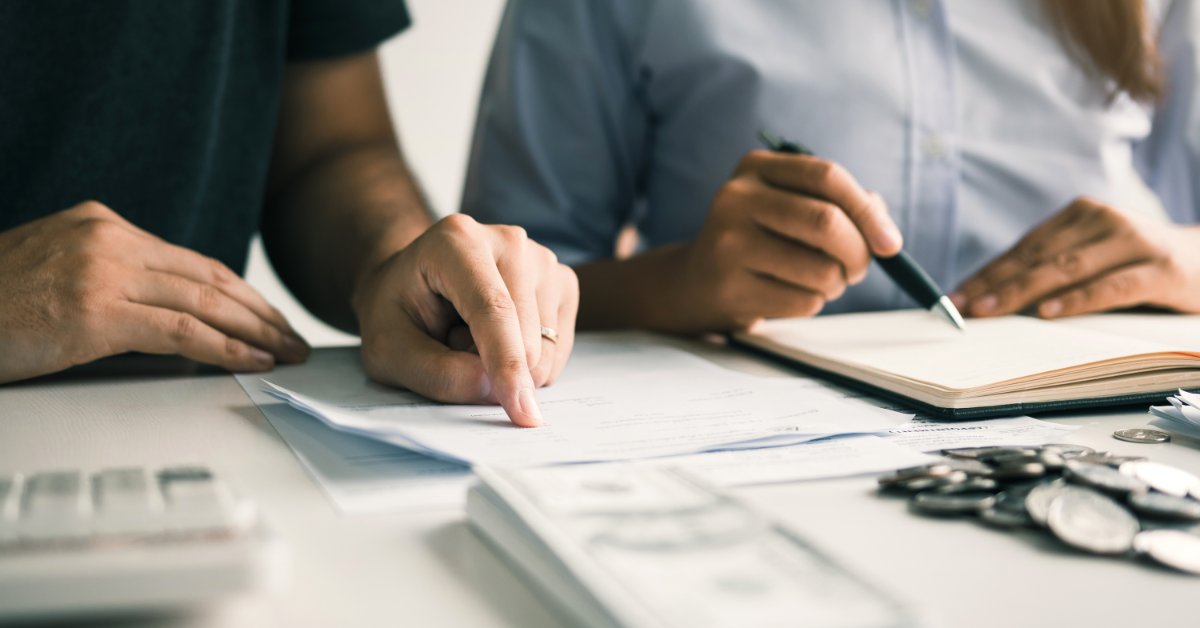 A man and a woman sitting behind a desk looking at multiple documents with a stack of cash, coins, and a calculator.