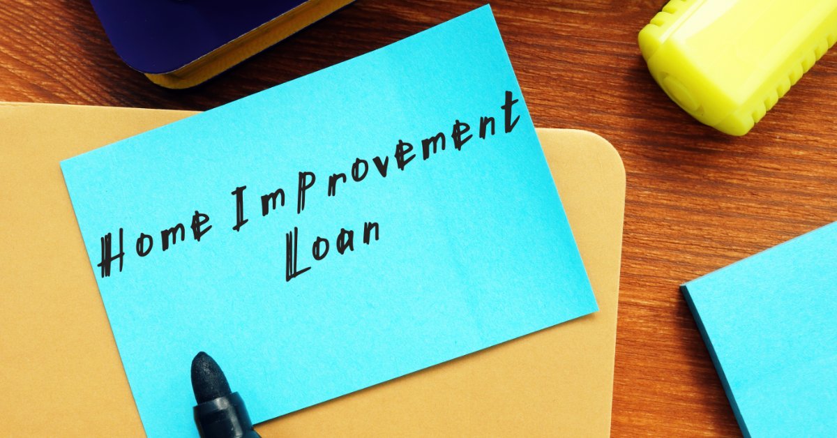An aerial view of a blue piece of paper displaying the words “home improvement loan” on a desk with office supplies.