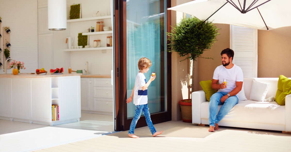 A boy walking out of a kitchen with sliding doors towards a man sitting on an outdoor couch under an umbrella.
