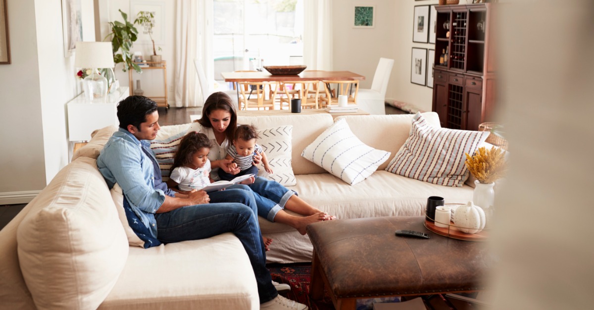 A man and a woman holding two small children while sitting on a sectional couch inside a large living room.