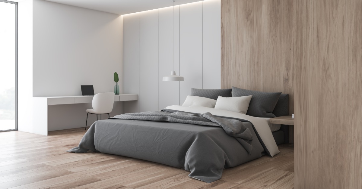 A room with a large bed in the center, wooden floors, and a white, large desk on the wall with a white chair.