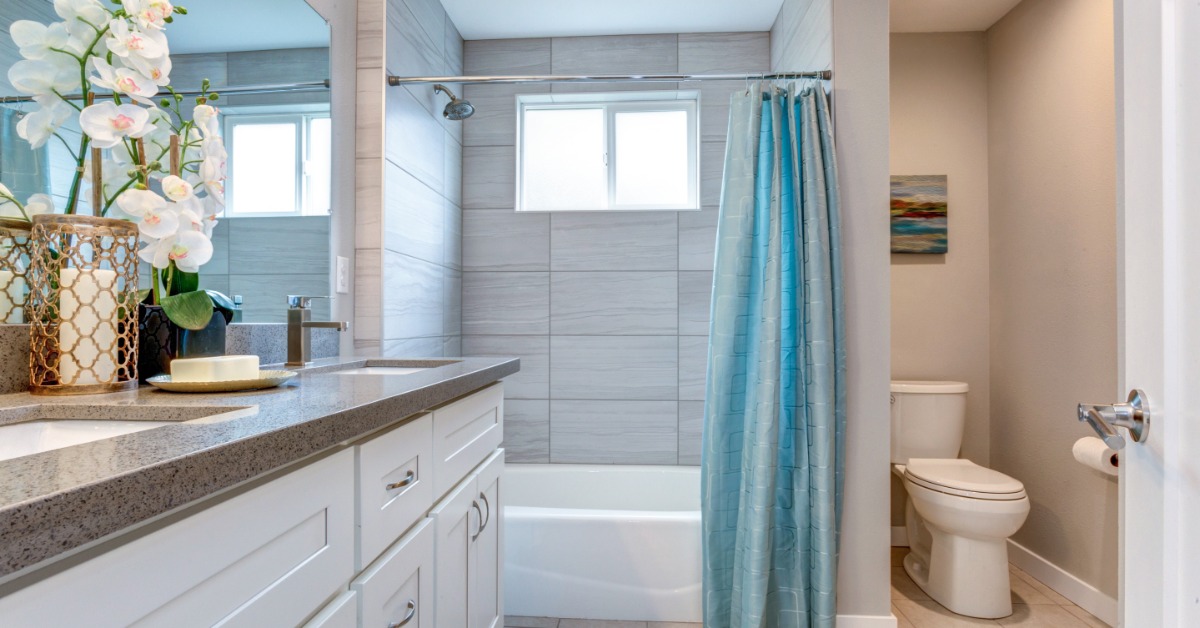 A bathroom with double sinks next to an orchid and a tub shower with a blue curtain next to a white toilet.