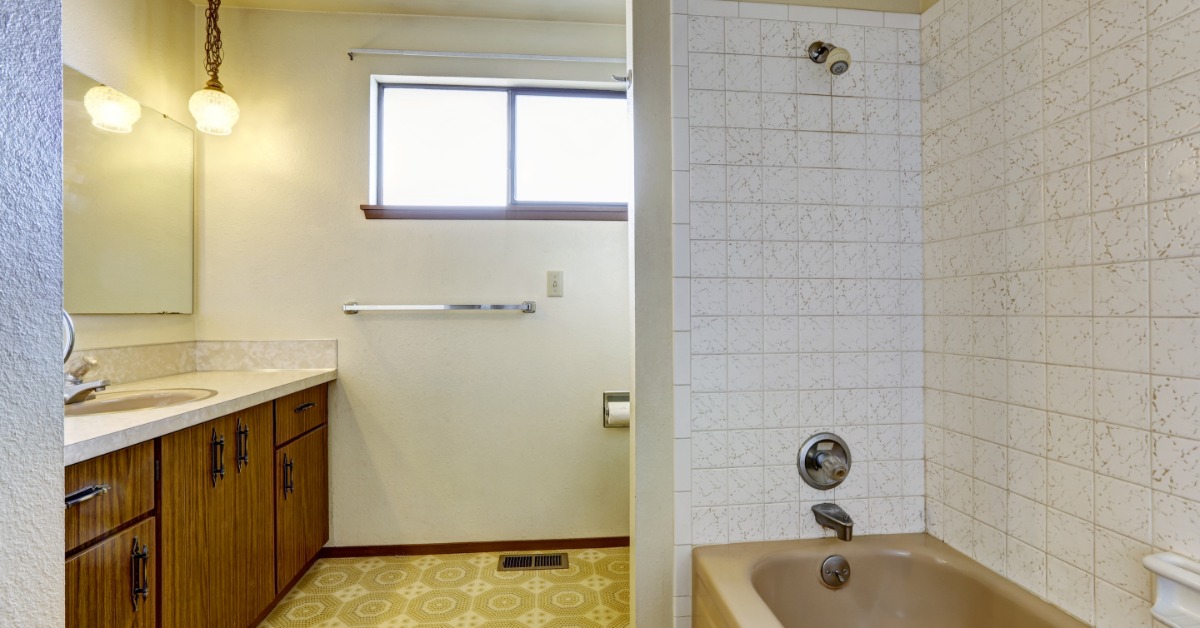 An old bathroom with yellow tiles on the floor, a pink bathtub, a small window, one lightbulb, and wooden cabinets.