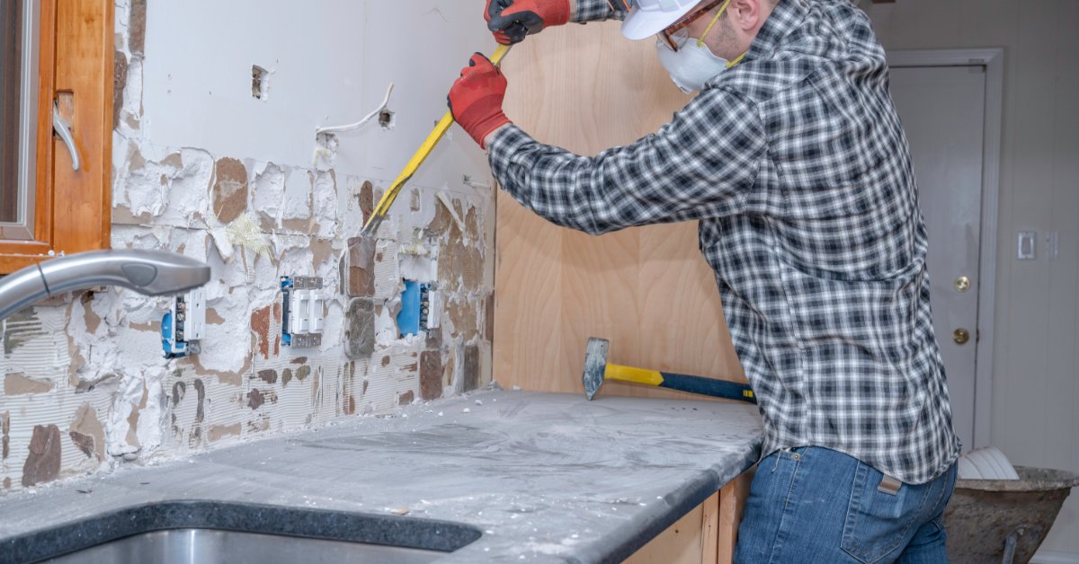 A man wearing a face mask and a helmet, using a metal bar to remove tiles from a kitchen wall above the counter.