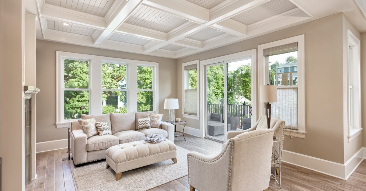 A room with a large couch, two cushioned chairs, a rug, hardwood floors, and large windows surrounded by greenery.