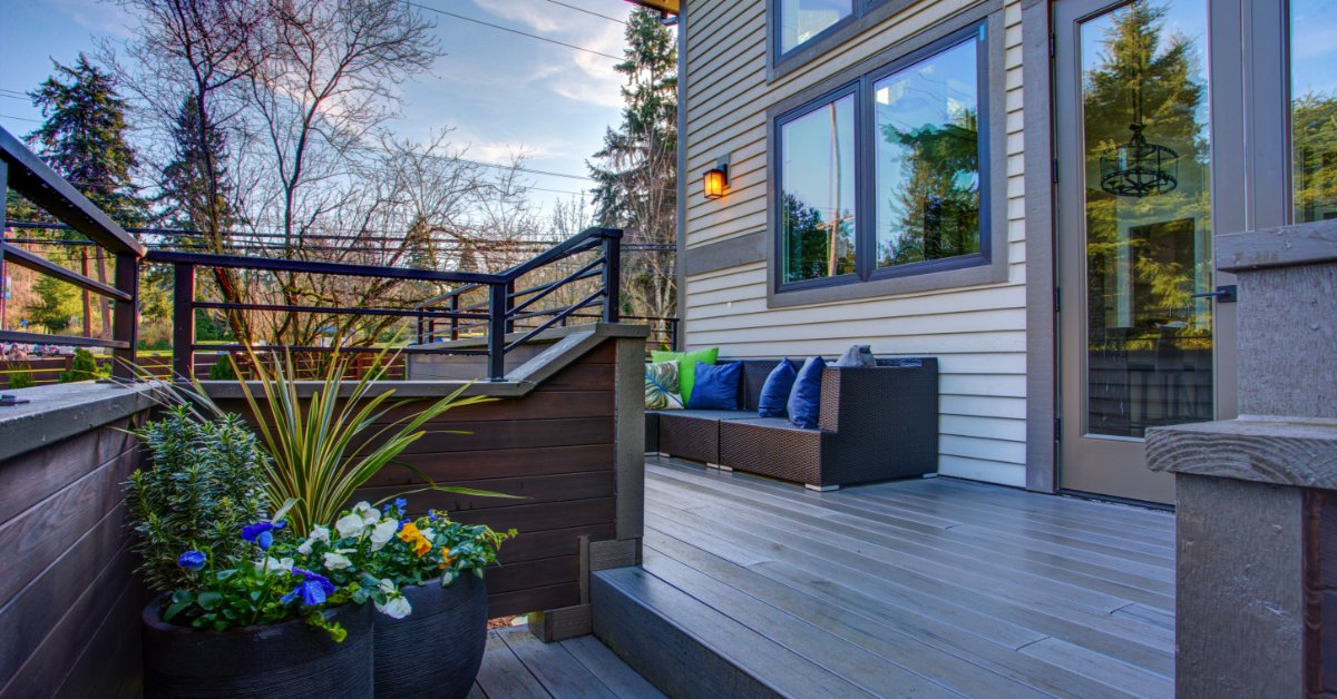 The exterior of a two-story home with paneled walls, a sectional outdoor couch, a handrail, and potted plants.