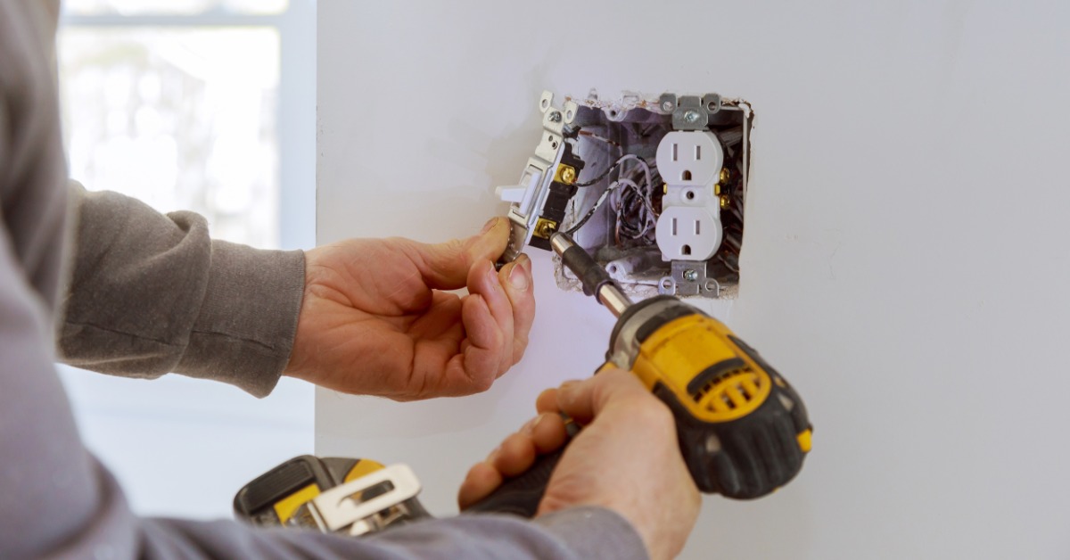 A close-up of two hands holding an electric screwdriver to install an electrical outlet and a switch on a white wall.