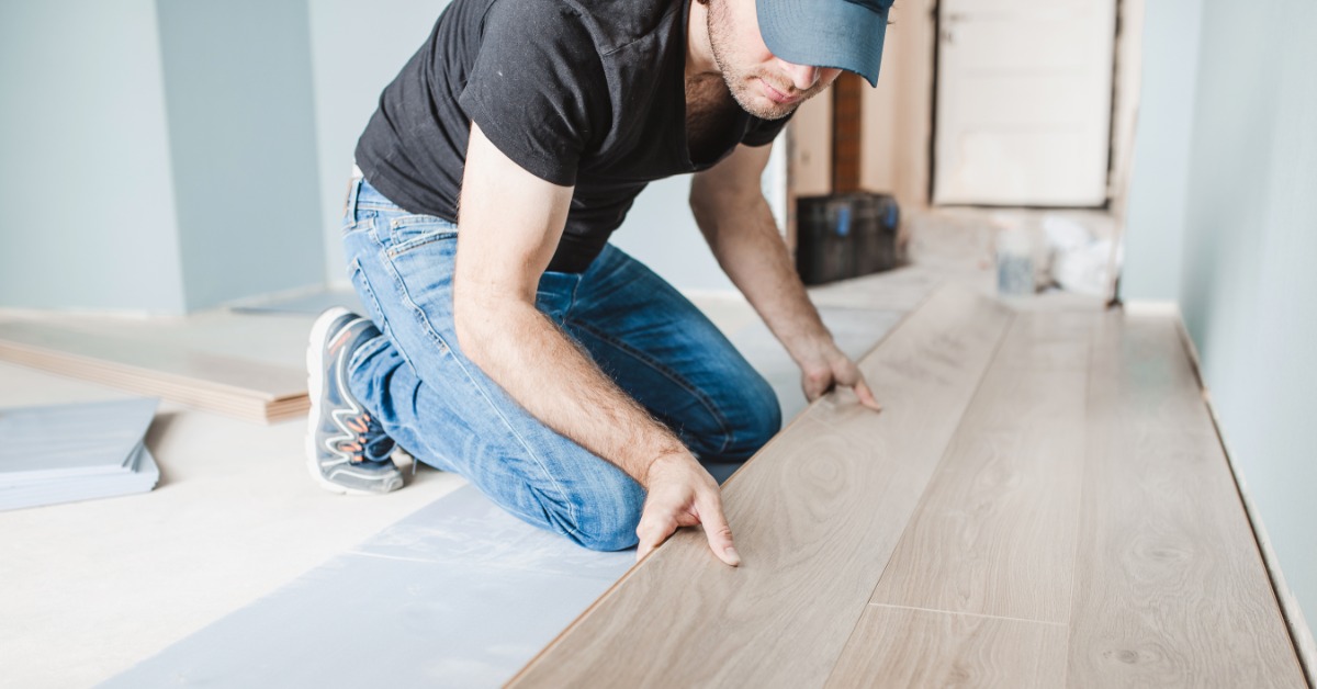 A man placing a long piece of laminated wood on a floor next to other pieces inside a room with blue walls.