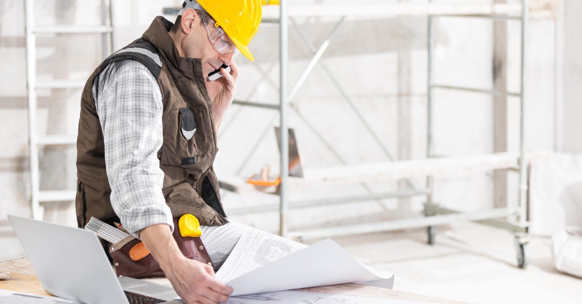 A man sitting on a desk, looking at a piece of paper while holding a smartphone to his ear in a room under construction.