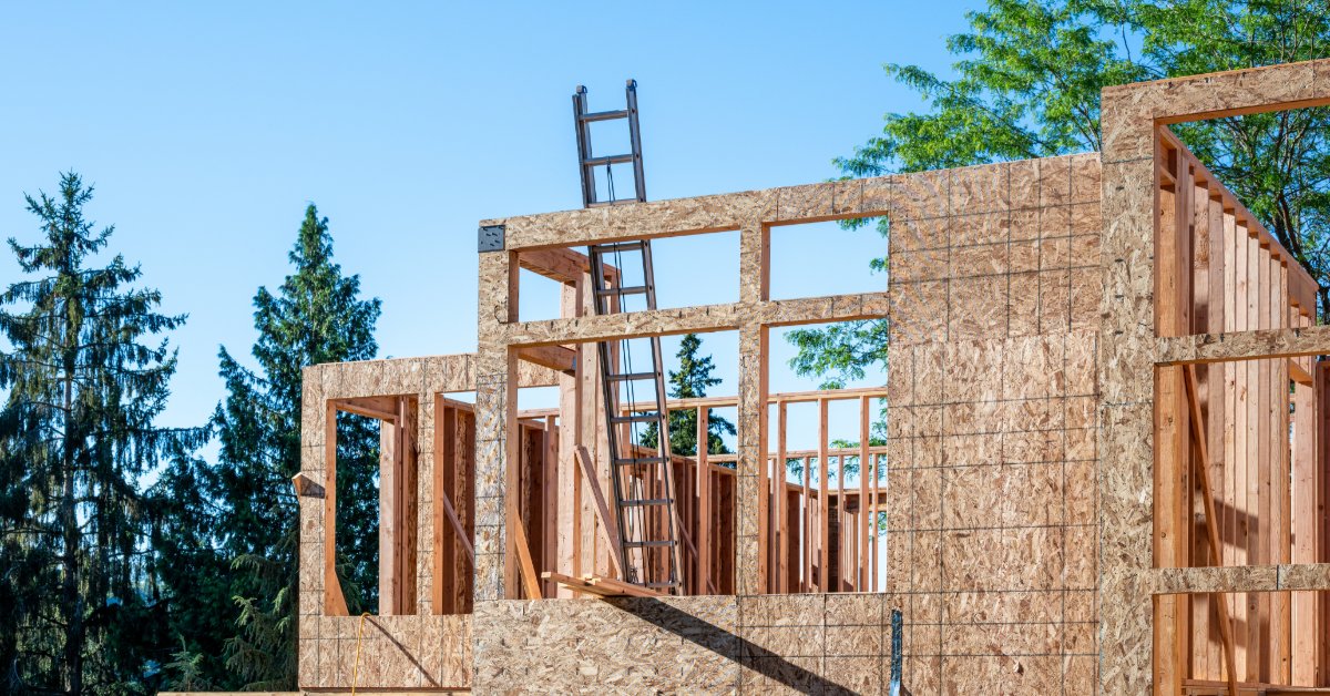 A worker wearing a safety vest and a helmet standing on top of the wooden skeleton of a house with a wooden beam.