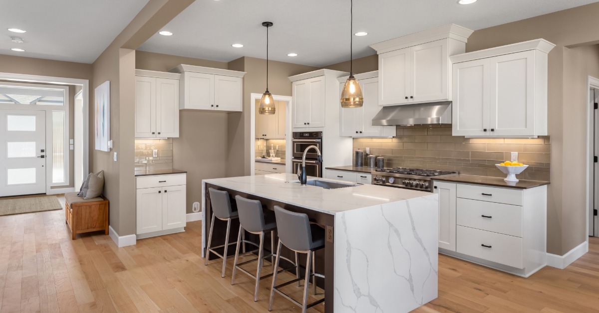 A large kitchen with wooden floors, an island with a marble counter, and white cabinets next to the main entrance.
