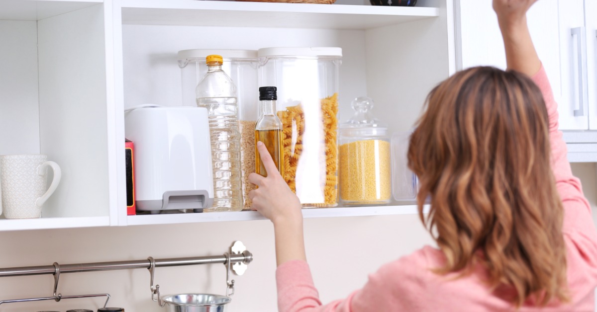 A woman wearing a pink blouse lifting the door of a white kitchen cabinet, placing a bottle of cooking oil inside.