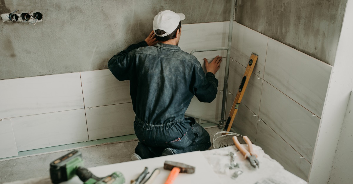 A worker wearing a grey uniform kneeling inside a bathroom under construction, placing a large tile on the wall.