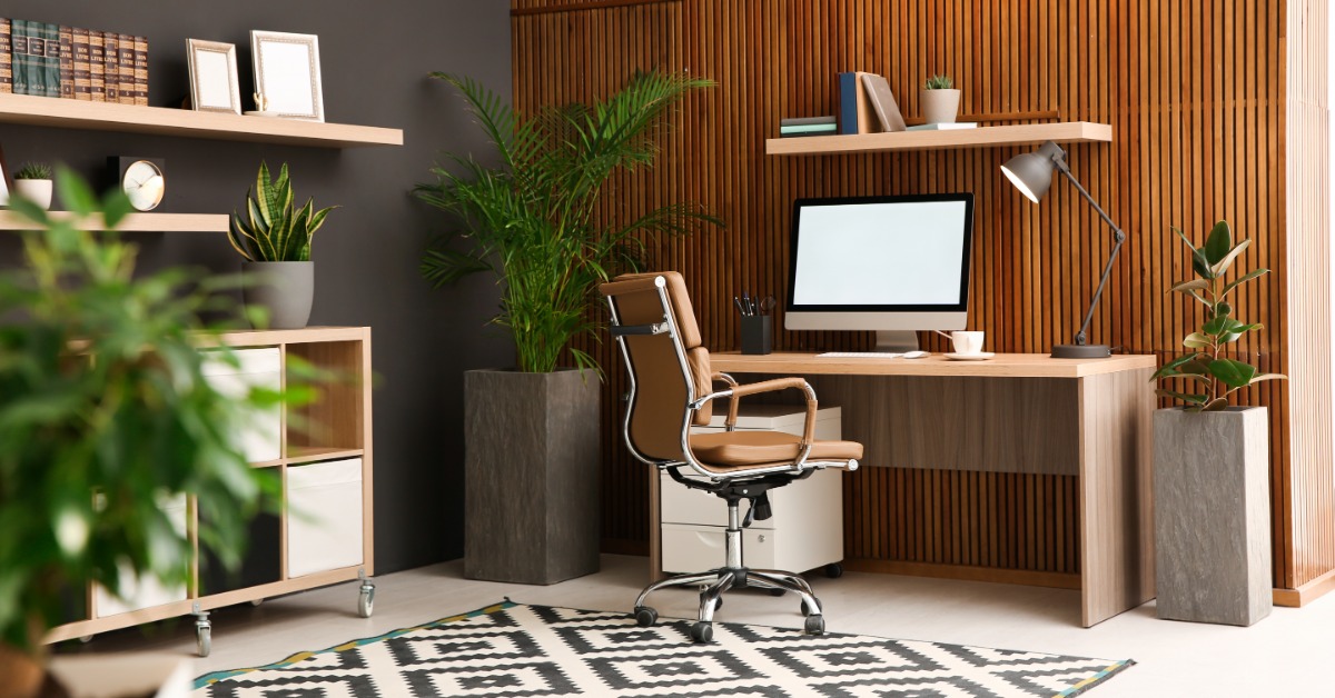 A room with a wooden wall, a large desk with a computer monitor, a chair, a bookshelf, and multiple green plants.