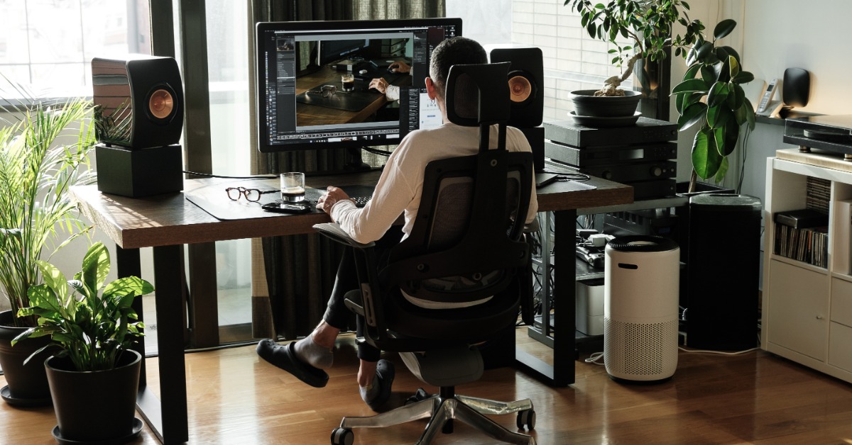 A person sitting on an ergonomic chair behind a desk with a large screen and speakers surrounded by small plants.