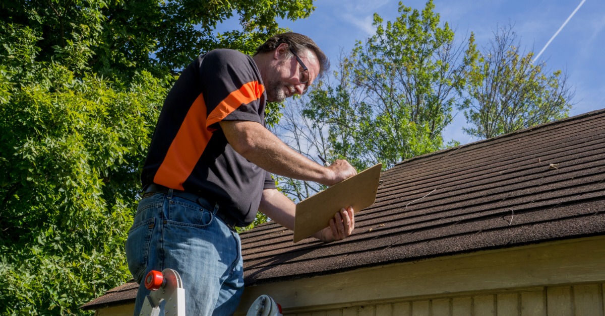 A man wearing a black shirt and glasses, standing on top of a ladder next to a roof, taking notes on a clipboard.
