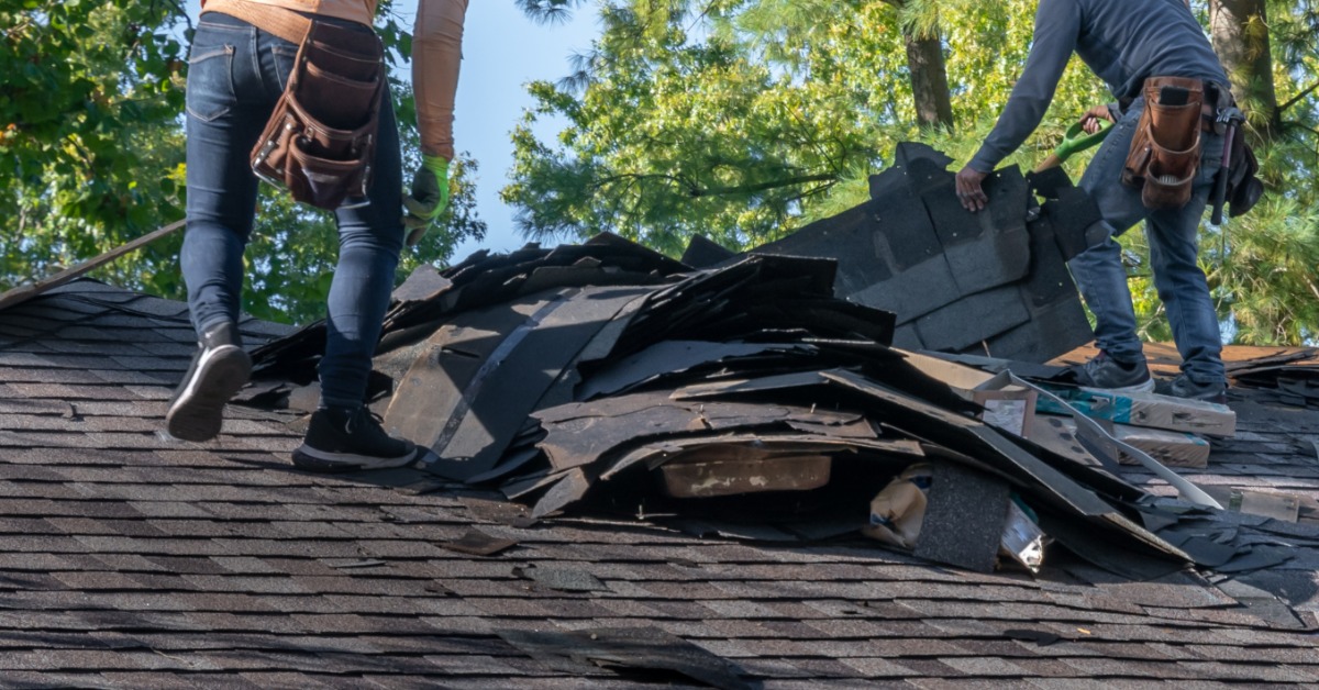 Two workers wearing jeans and leather tool belts removing shingles from a home’s roof, putting them in a pile.